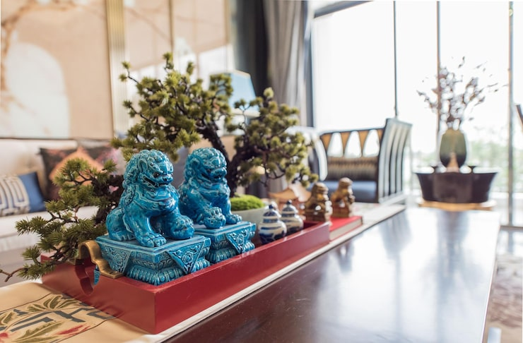 a living room corner with blue foo dogs, a bonsai tree, small decorative items on a red tray, and a stylish background with a sofa and large windows