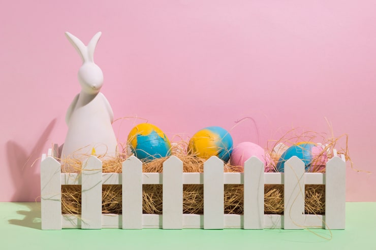 easter decorations with a white rabbit figurine and colorful eggs in a white picket fence box against a pink background